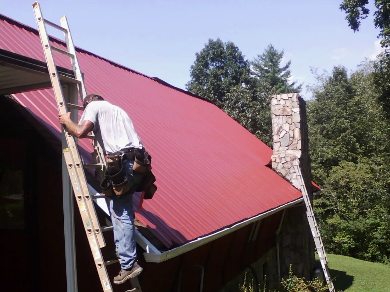 Roofer on a ladder beside a red metal roof and stone chimney — Soffit & Fascia Repair by Roofing Brothers in Ballantine